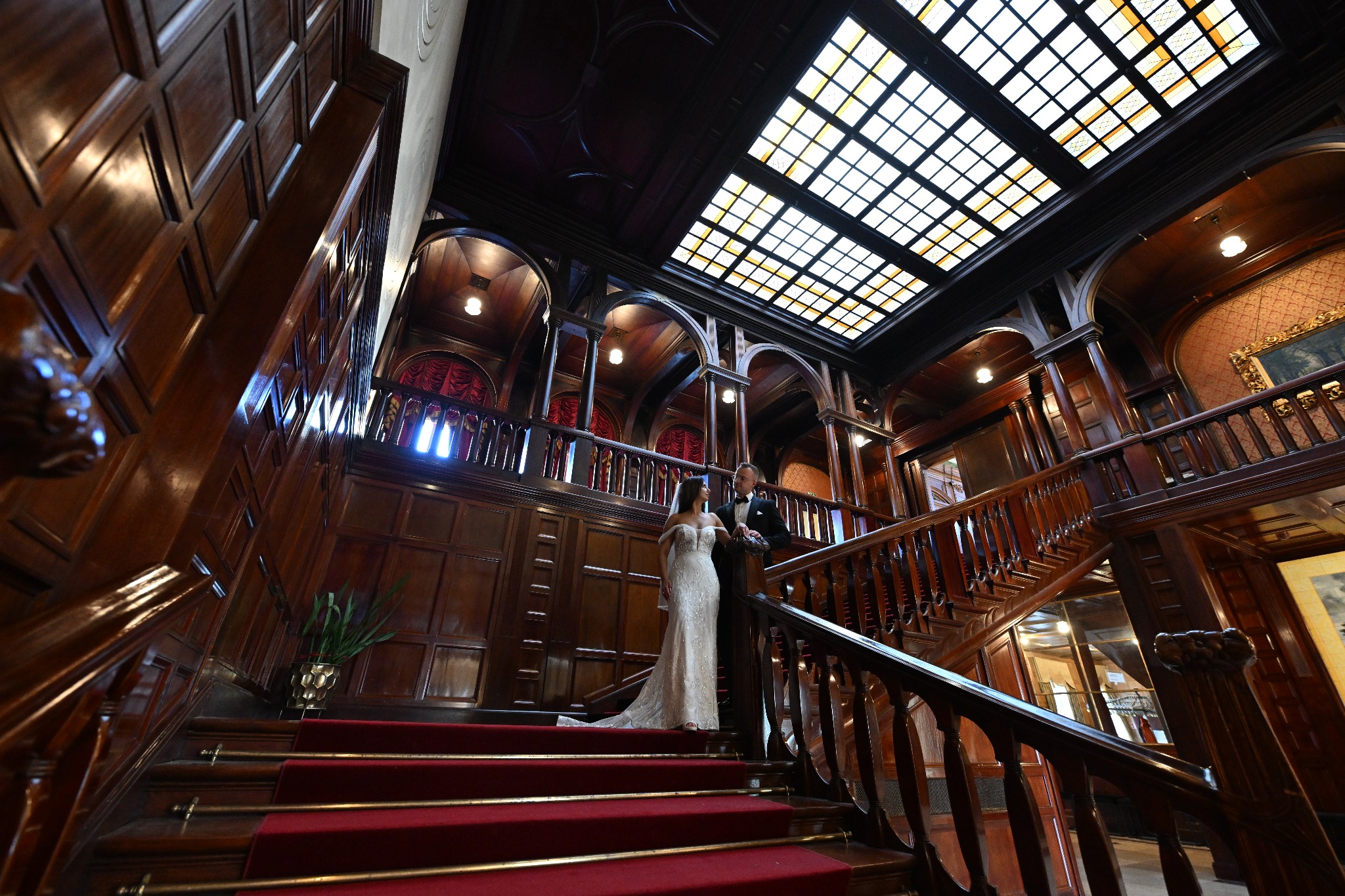 Para młoda w eleganckich strojach stoi na czerwonych schodach w bogato zdobionym, drewnianym wnętrzu z przeszklonym sufitem.