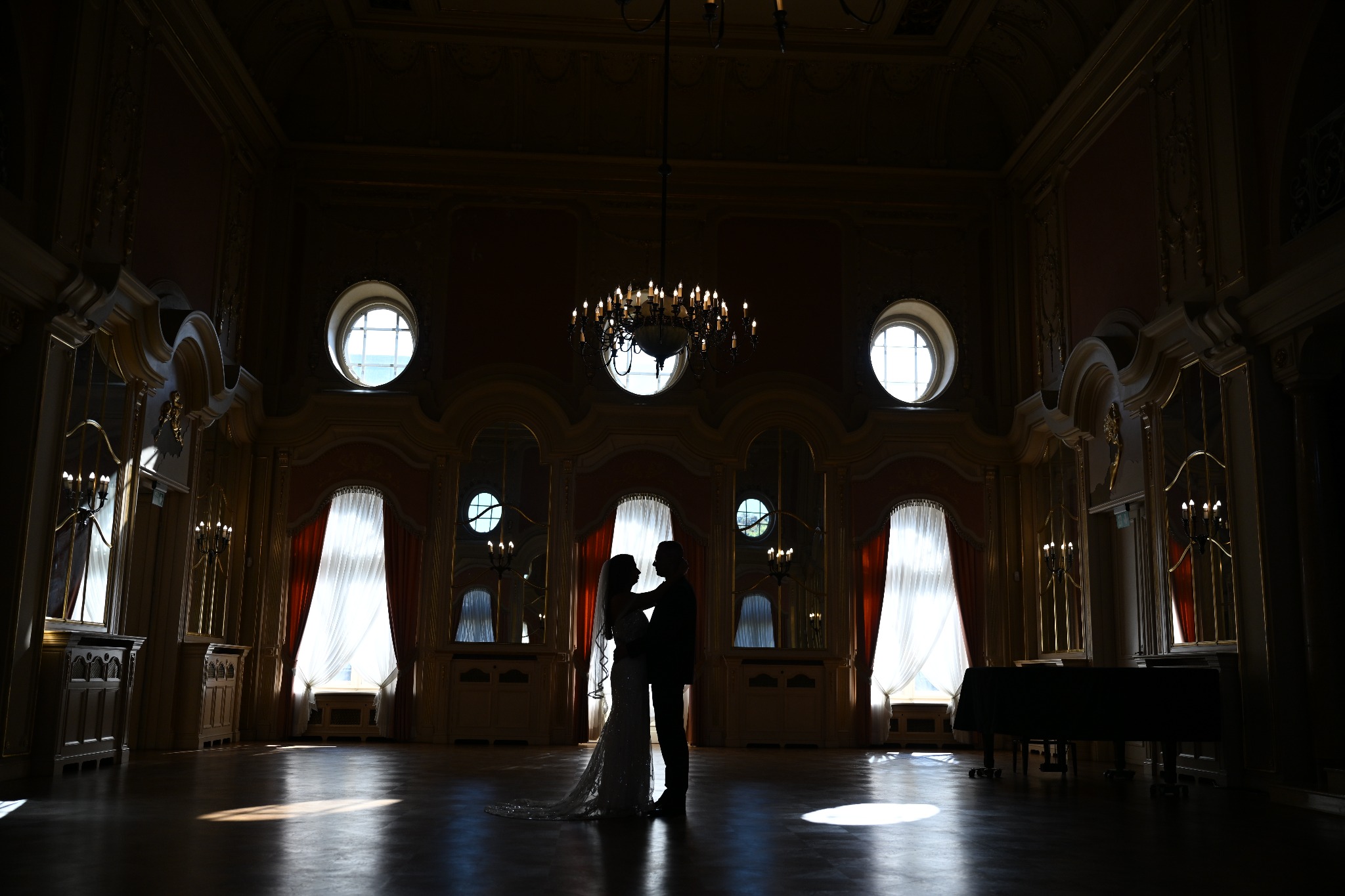 Sylwetki pary młodej w objęciach na tle bogato zdobionej sali balowej z żyrandolem i dużymi oknami.