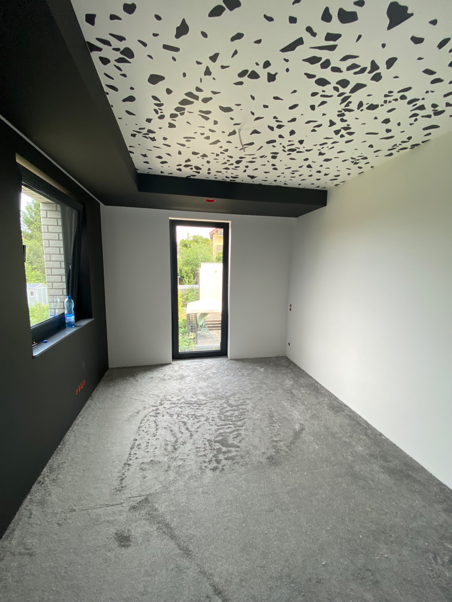 Pokój po remoncie: pomalowane ściany na biało i szaro, sufit z wzorem w czarne plamy, podłoga przygotowana pod układanie parkietu, widoczne okno i drzwi.