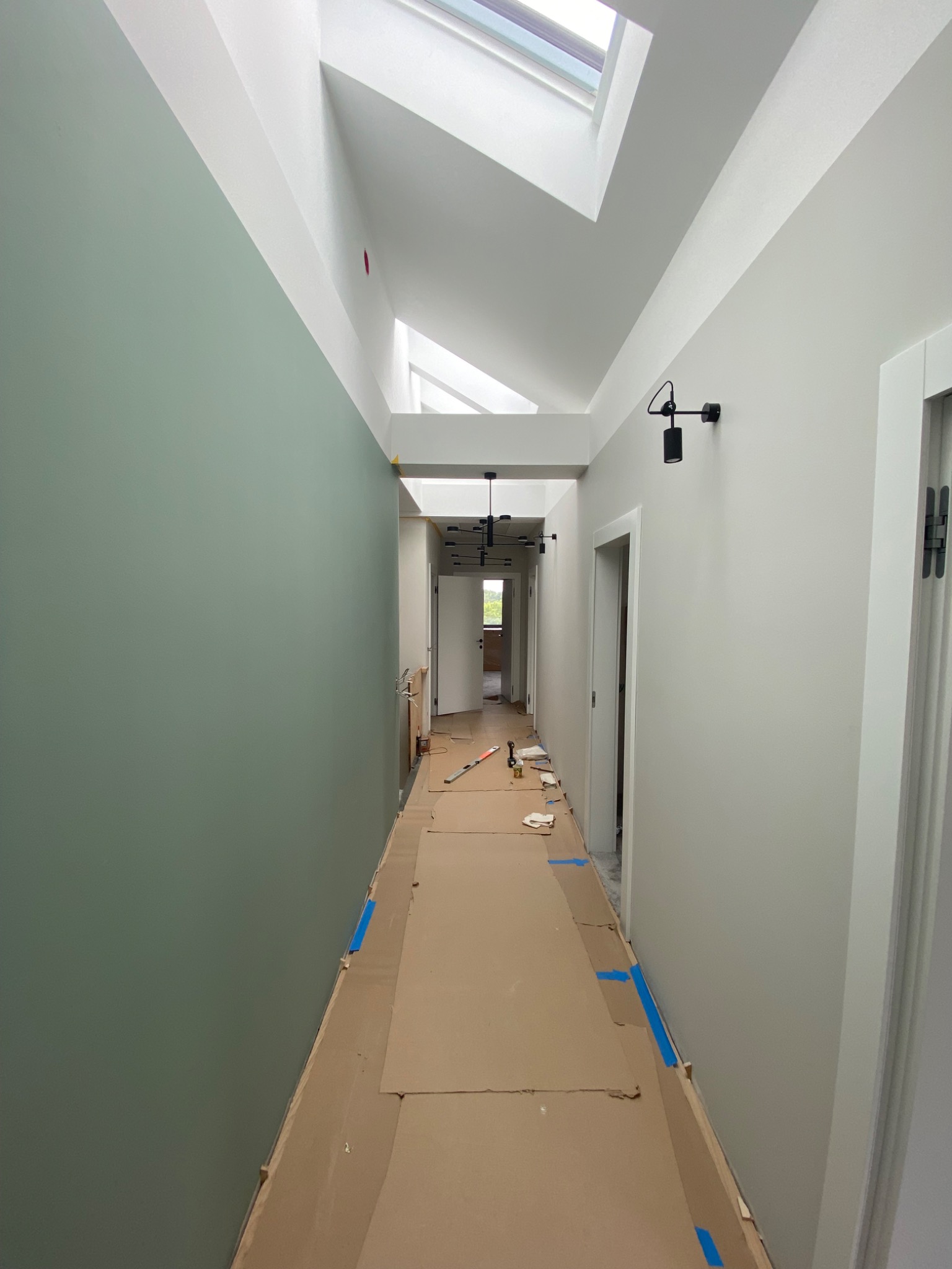 Korytarz w trakcie prac wykończeniowych: ściany pomalowane w dwóch odcieniach, zielonym i białym, zabezpieczona podłoga kartonem, widoczne narzędzia malarskie i oświetlenie sufitowe.