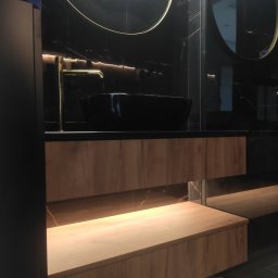 Meble KADE - Nowoczesna łazienka z czarnymi marmurowymi ścianami, okrągłymi lustrami z podświetleniem LED, drewnianą szafką pod umywalką z oświetleniem od dołu i złotą armaturą.
