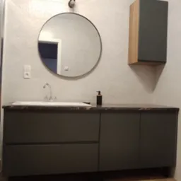 Nowoczesna łazienka z ciemnoszarą szafką pod umywalkę z blatem imitującym kamień, okrągłym lustrem i szafką wiszącą z drewnianymi bokami na ścianie z trójkątną fakturą.