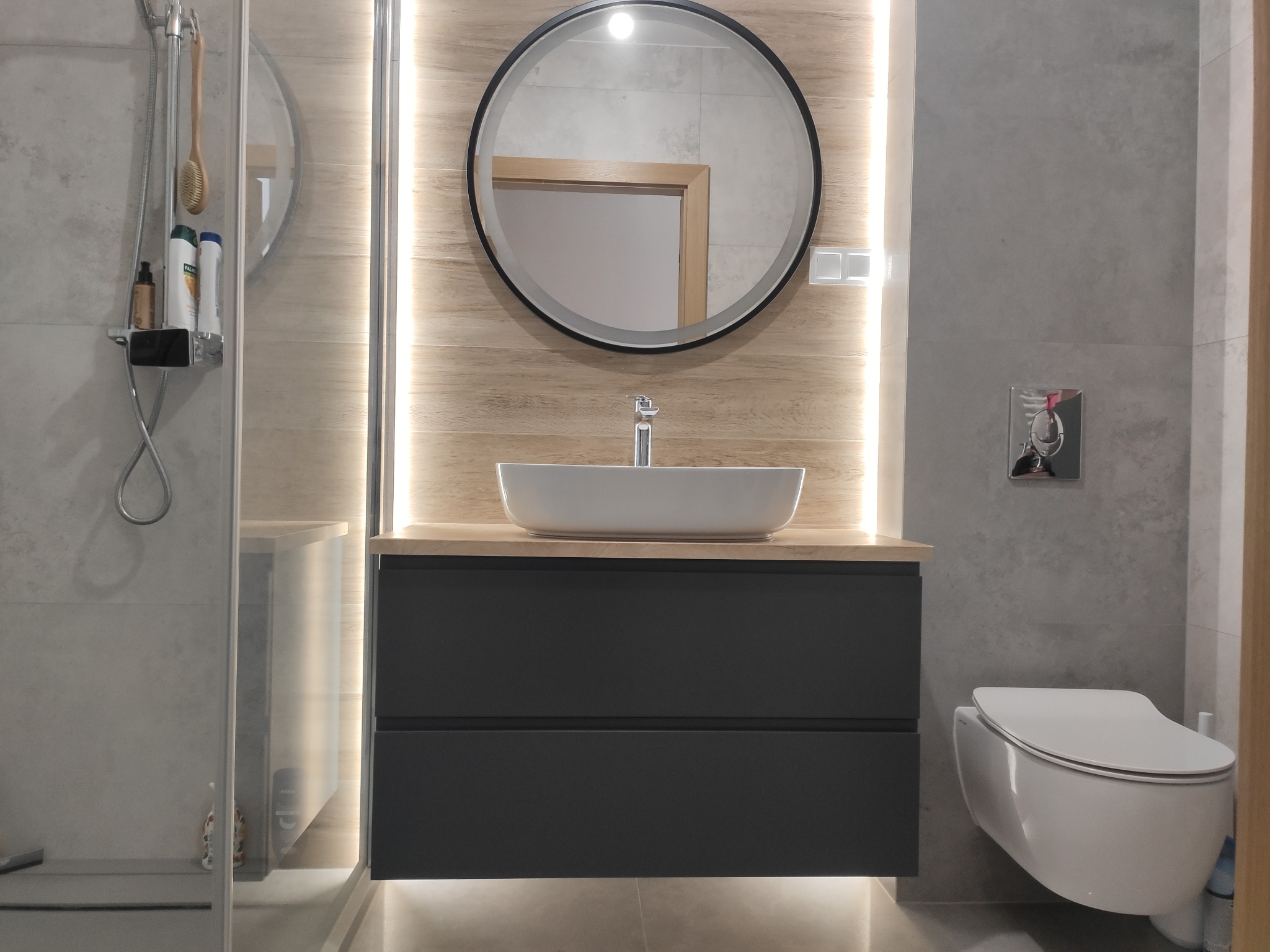 Nowoczesna łazienka z szarą armaturą, podświetlanym lustrem okrągłym i drewnianym blatem pod umywalką nablatową.