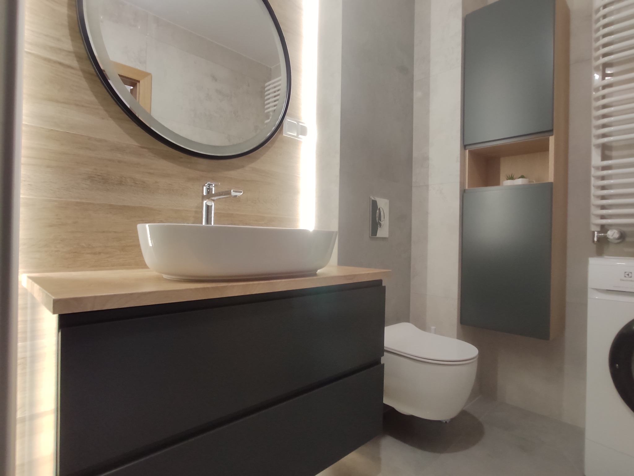 Nowoczesna łazienka z umywalką nablatową na czarnej szafce z szufladami, okrągłym lustrem z podświetleniem, wysoką szafką wiszącą w kolorze grafitowym i grzejnikiem drabinkowym.