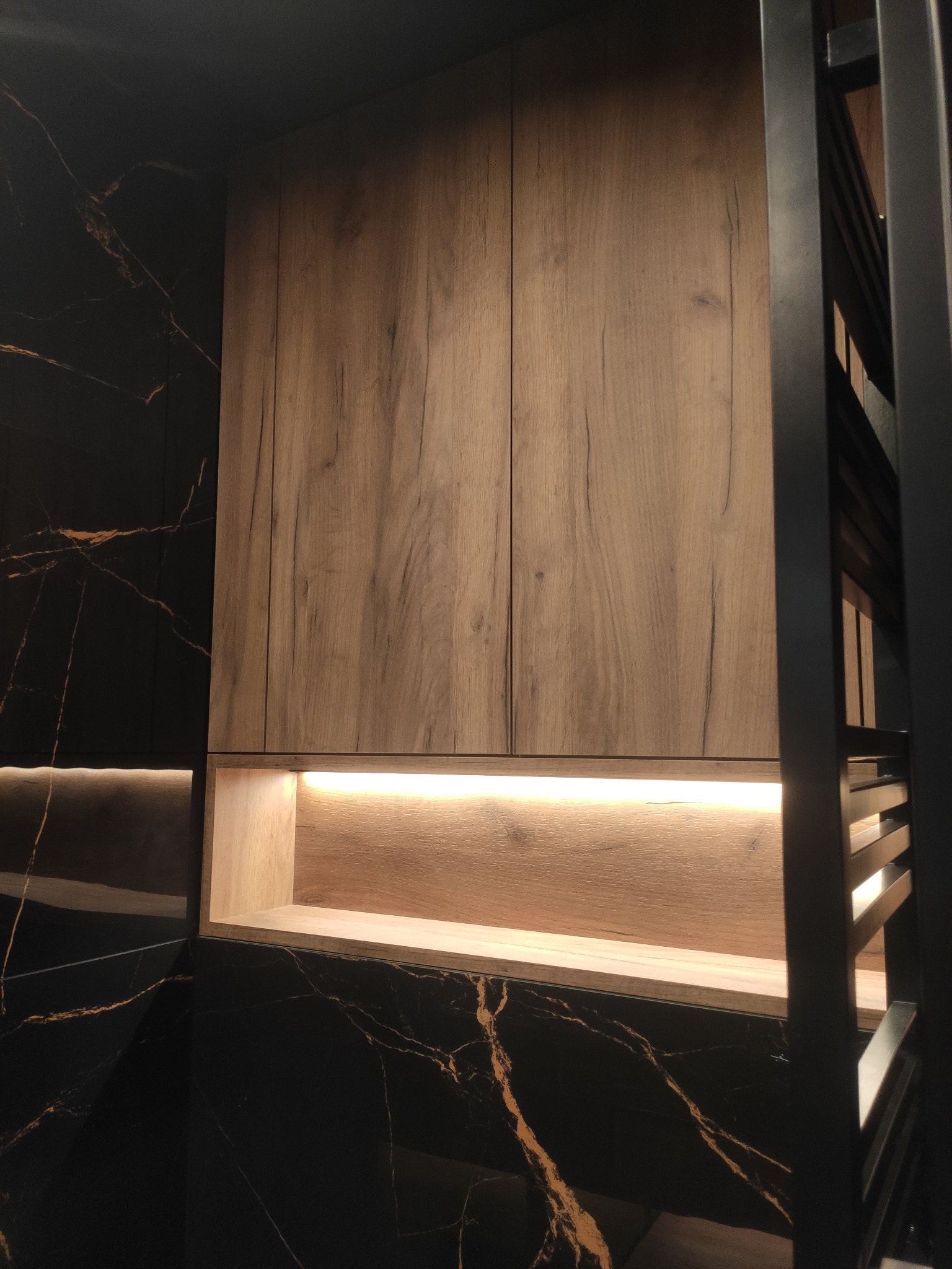 Drewniana szafka łazienkowa z oświetleniem LED, zamontowana na ścianie z czarnego marmuru z złotymi żyłami, obok grzejnika drabinkowego.