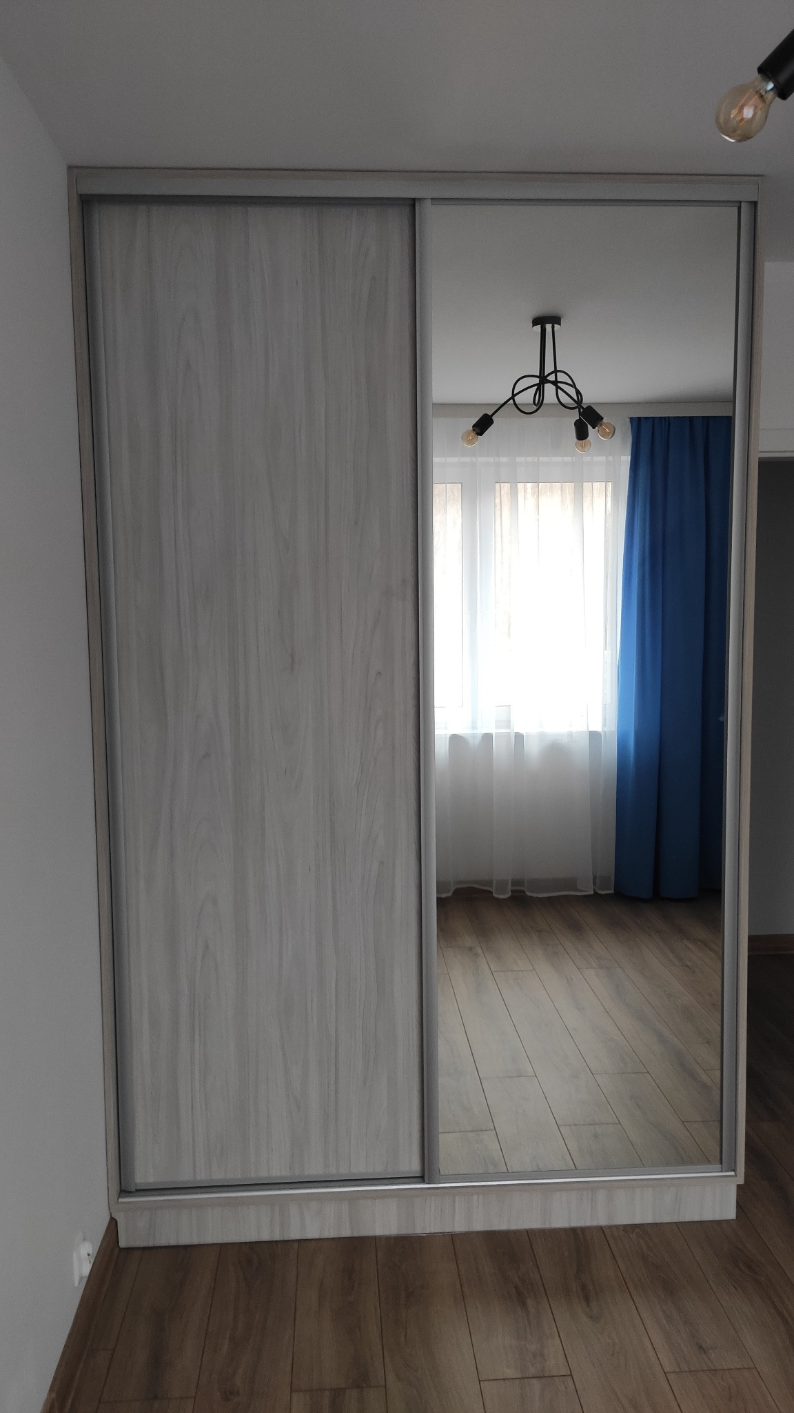 Szafa z przesuwnymi drzwiami, jedno skrzydło z płyty imitującej drewno, drugie lustrzane, odbijające wnętrze pokoju z lampą i zasłonami.