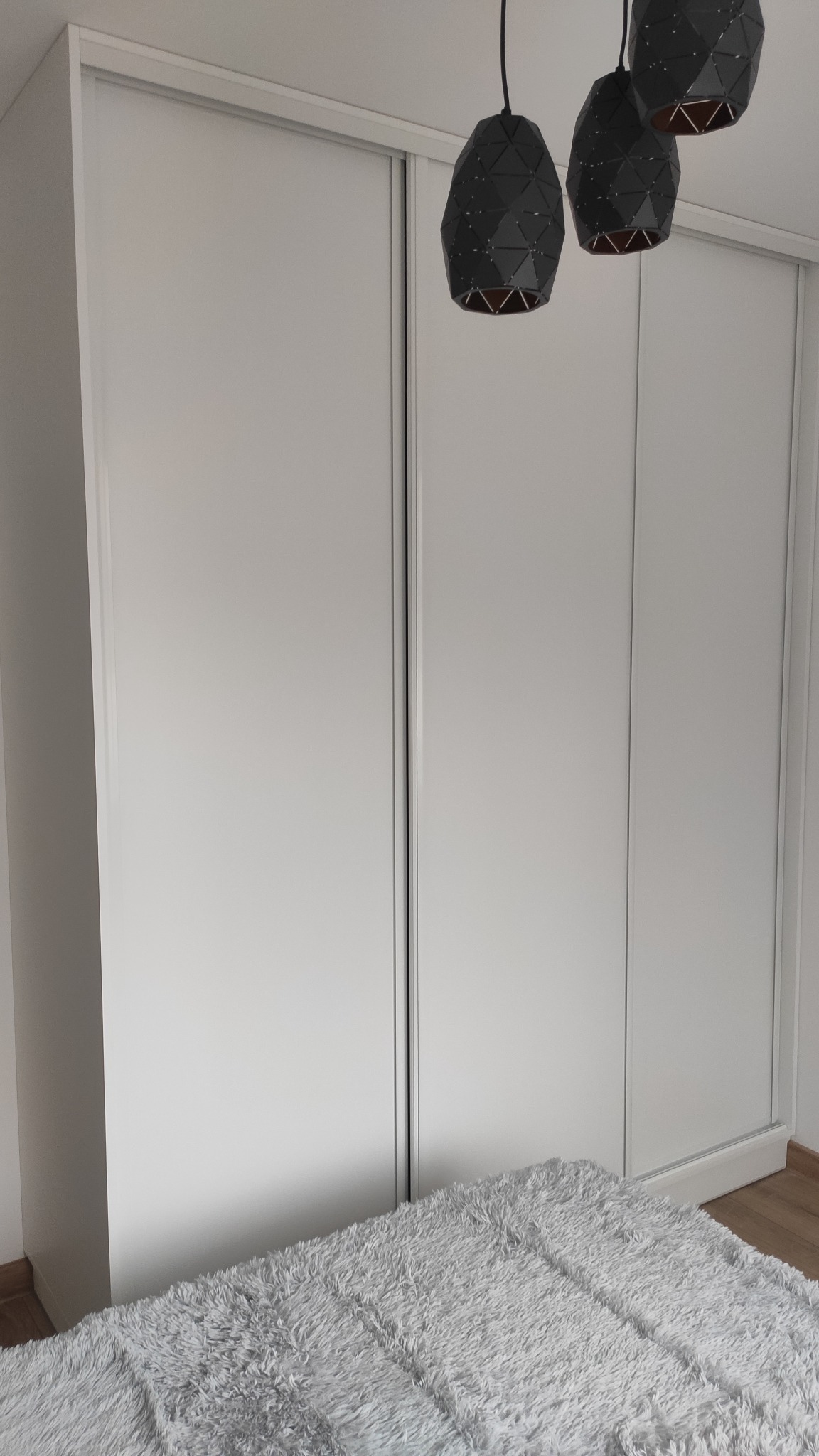Trzydrzwiowa szafa przesuwna z białymi frontami, oświetlona trzema geometrycznymi lampami wiszącymi, na pierwszym planie puszysty, szary dywan.