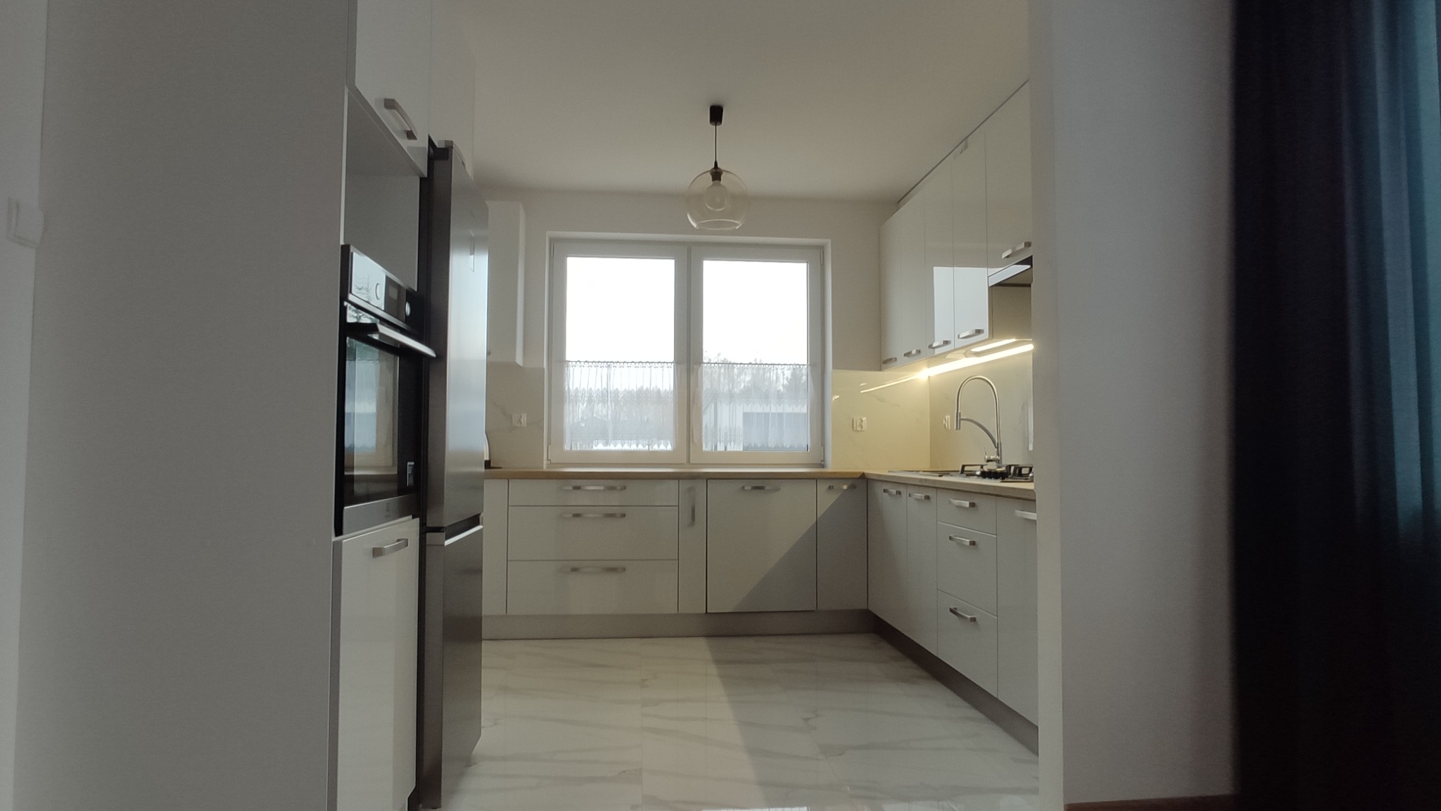 Jasna kuchnia z białymi szafkami i marmurową podłogą, oświetlona lampą wiszącą, widok zza ściany.