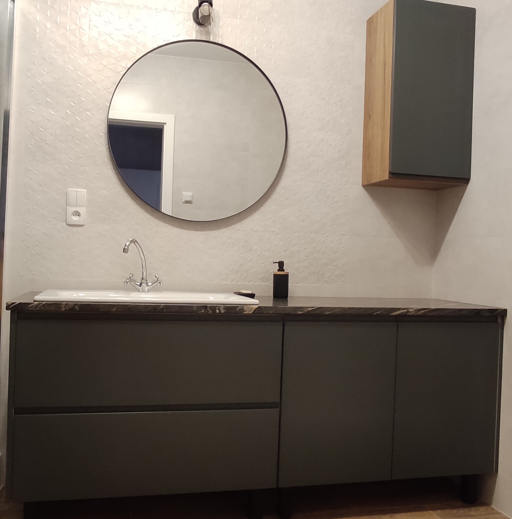 Nowoczesna łazienka z ciemnoszarą szafką pod umywalkę z blatem imitującym kamień, okrągłym lustrem i szafką wiszącą z drewnianymi bokami na ścianie z trójkątną fakturą.