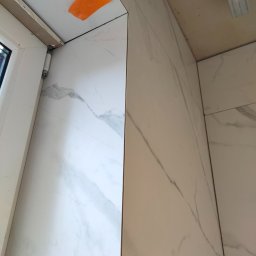 Dawid Szumlański - Fragment ściany łazienki wyłożonej białymi płytkami imitującymi marmur, widoczne narożniki i okno, zabezpieczone pomarańczową taśmą malarską, sufit w trakcie montażu.