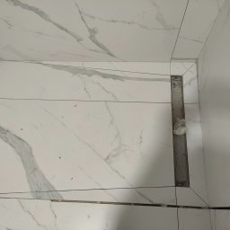 Dawid Szumlański - Białe płytki imitujące marmur, ułożone pod prysznicem, z widocznym odpływem liniowym. Płytki ułożone z precyzją, z zachowaniem spójności wzoru.