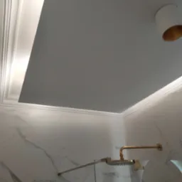 Wykończona łazienka z marmurowymi płytkami, złotą armaturą prysznicową i podwieszanym sufitem z ozdobną sztukaterią i oświetleniem punktowym.