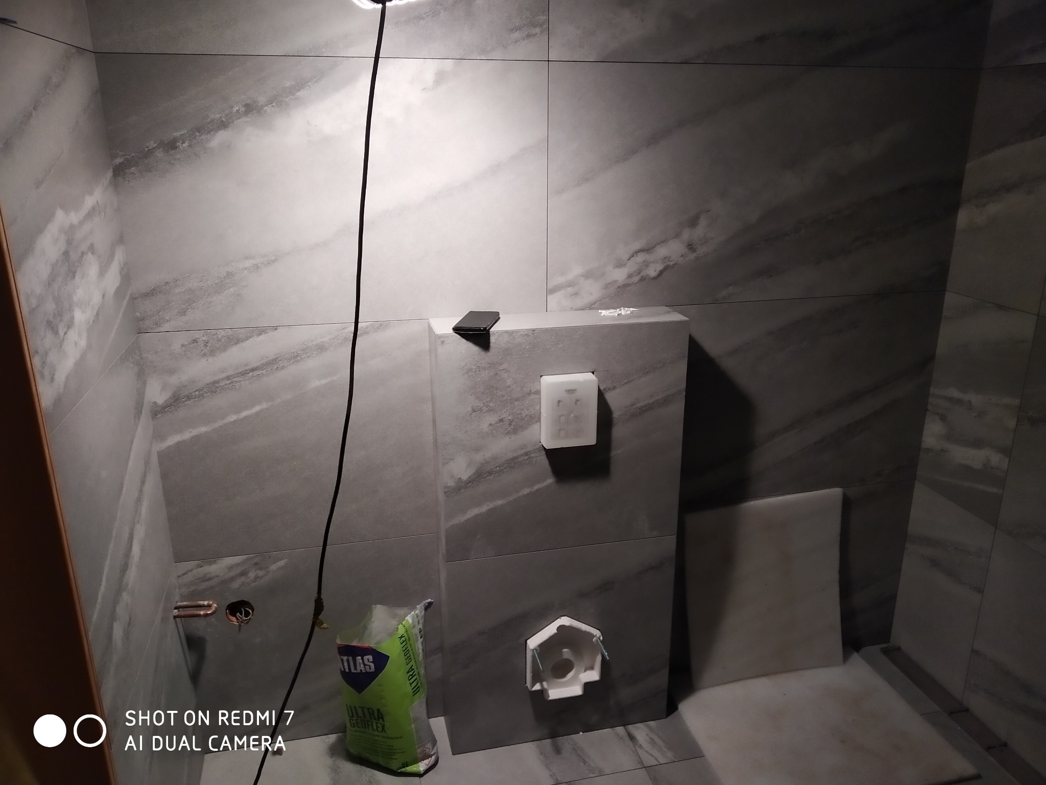 Fragment łazienki w trakcie prac, z szarymi płytkami imitującymi marmur, widoczna instalacja podtynkowa, worek z klejem Atlas Ultra Geoflex i telefon na zabudowie stelaża podtynkowego.
