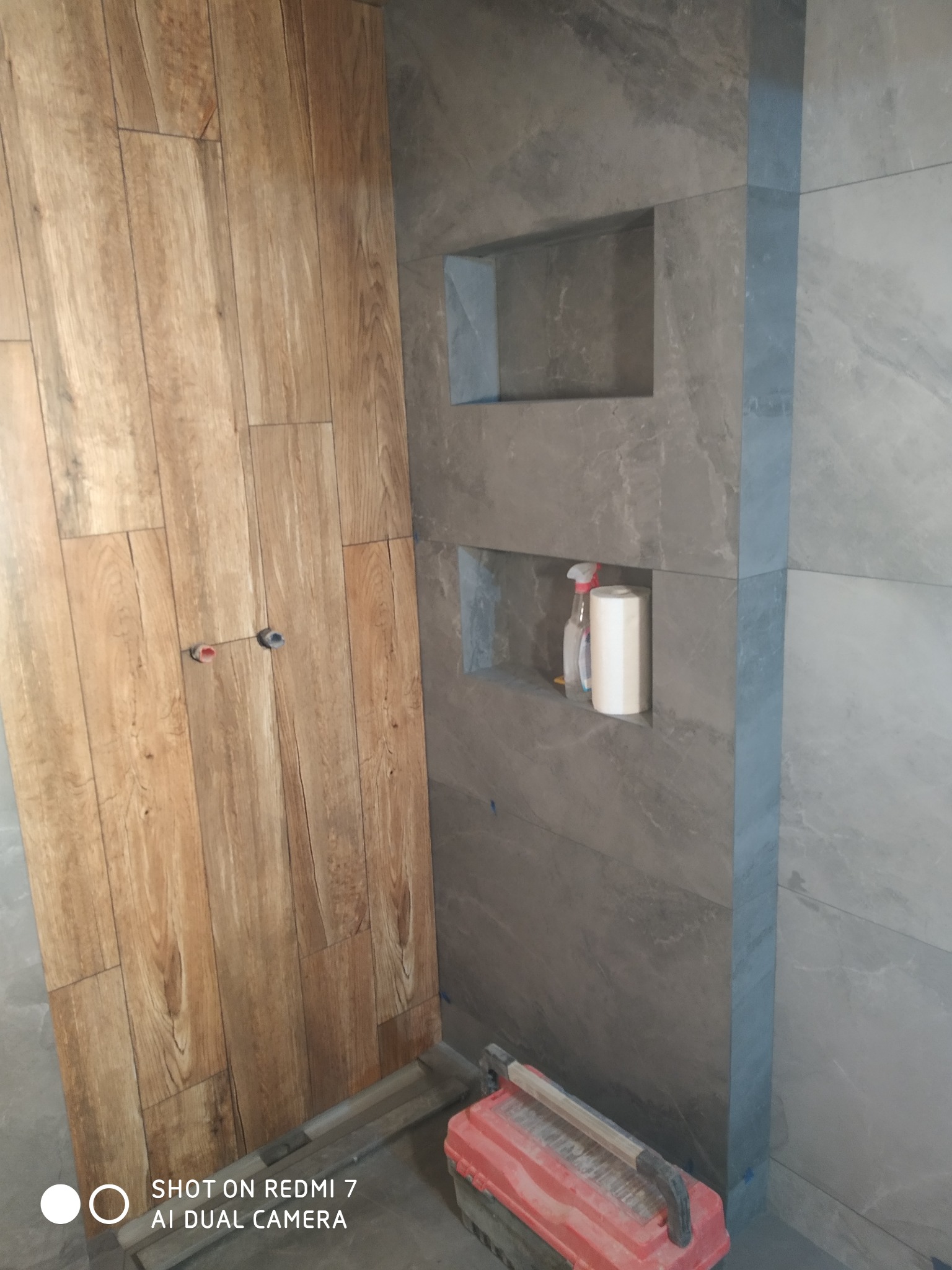 Fragment łazienki w trakcie wykańczania: drewnopodobne płytki na ścianie z widocznymi przyłączami, szare płytki imitujące kamień z wnękami, w jednej z nich papier toaletowy i butelka z atomizerem...