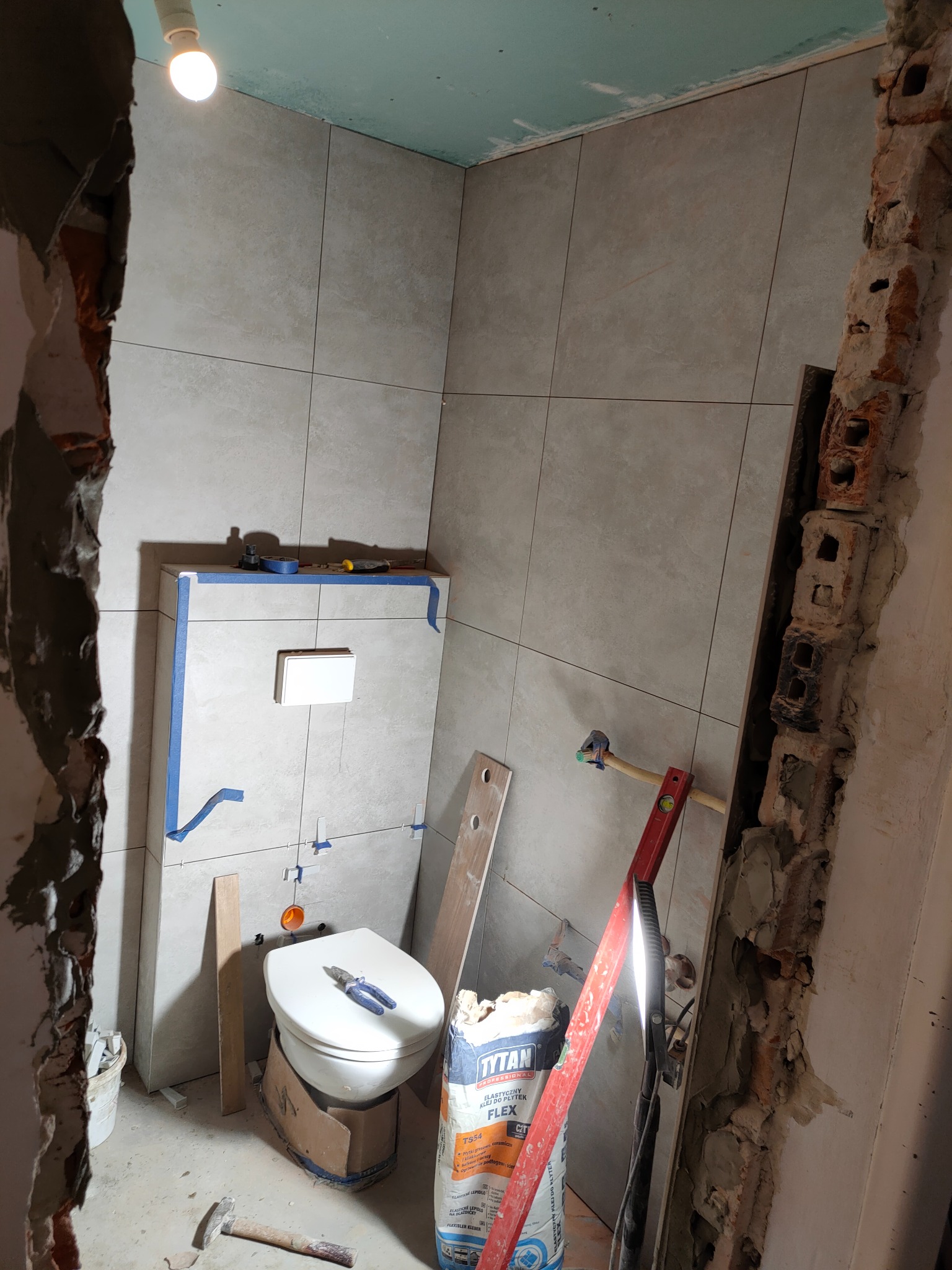 Remont łazienki w trakcie: ułożone szare płytki na ścianach, częściowo zamontowana toaleta z narzędziami na desce, worek zaprawy Tytan Flex, poziomica i odsłonięta cegła.