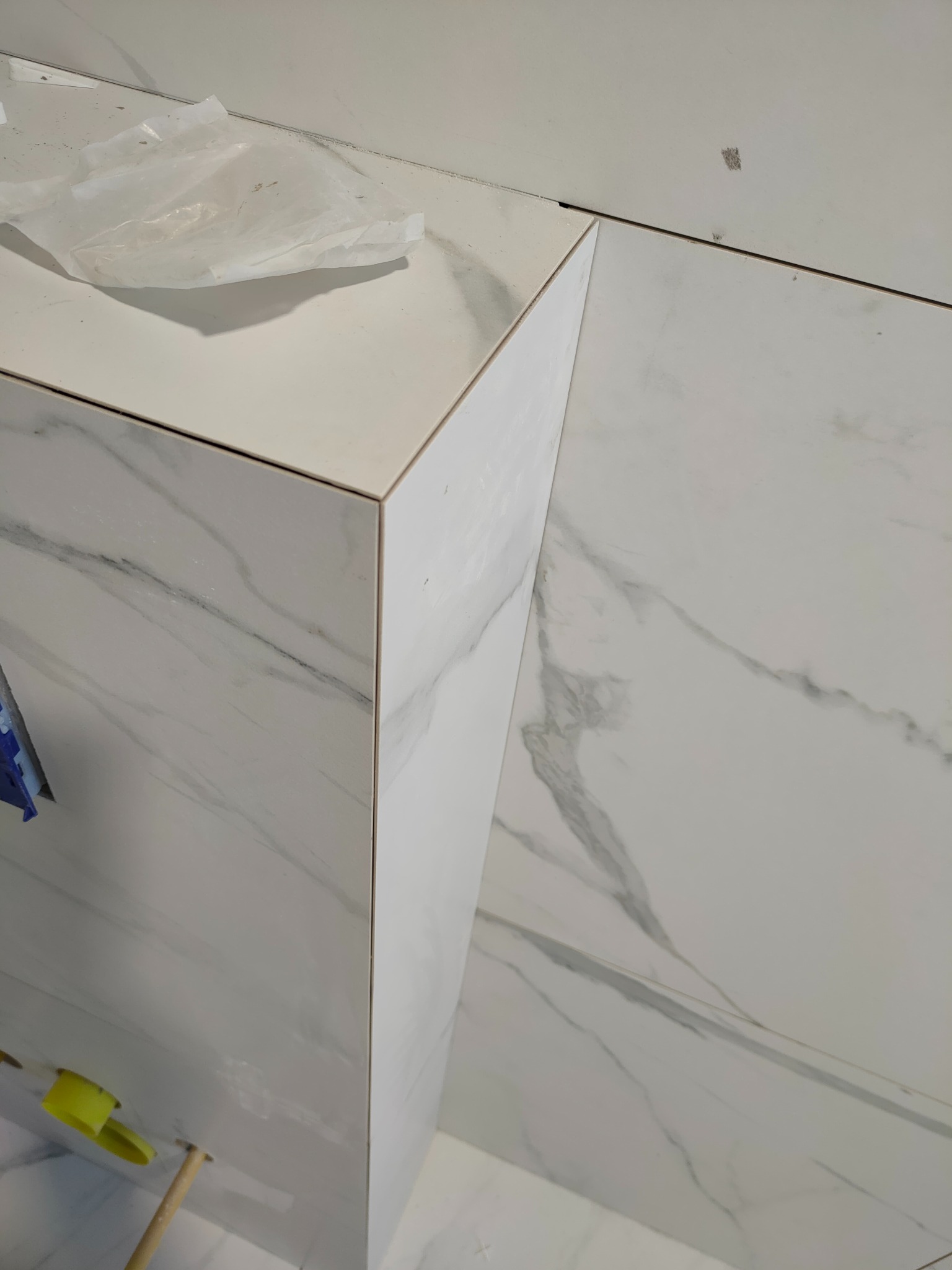 Fragment wykończonej ściany z białych płytek imitujących marmur, widoczny fragment żółtego poziomowania i resztki folii ochronnej na blacie.