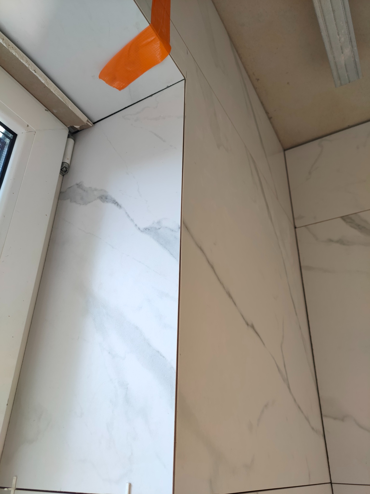 Fragment ściany łazienki wyłożonej białymi płytkami imitującymi marmur, widoczne narożniki i okno, zabezpieczone pomarańczową taśmą malarską, sufit w trakcie montażu.