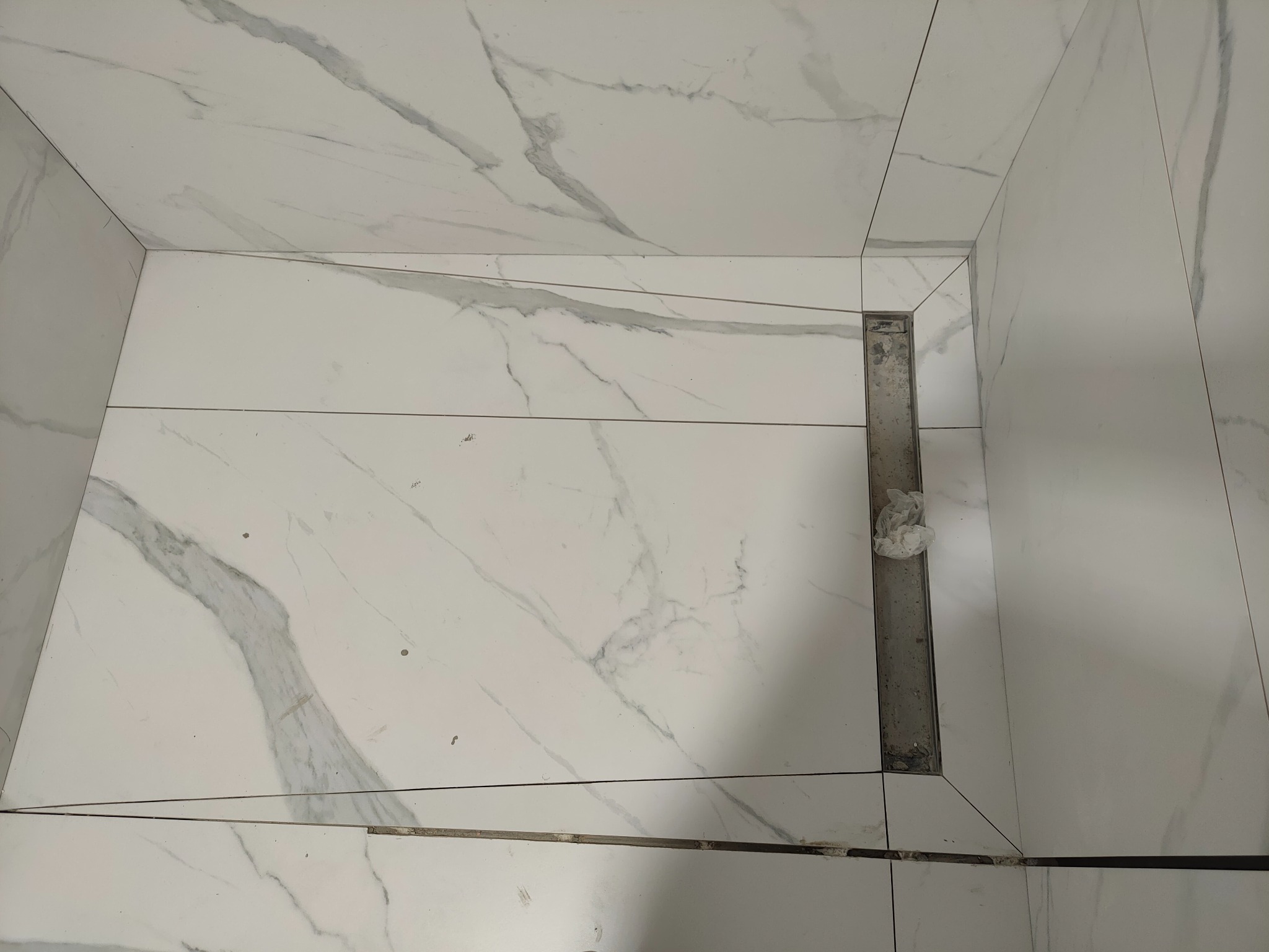 Białe płytki imitujące marmur, ułożone pod prysznicem, z widocznym odpływem liniowym. Płytki ułożone z precyzją, z zachowaniem spójności wzoru.