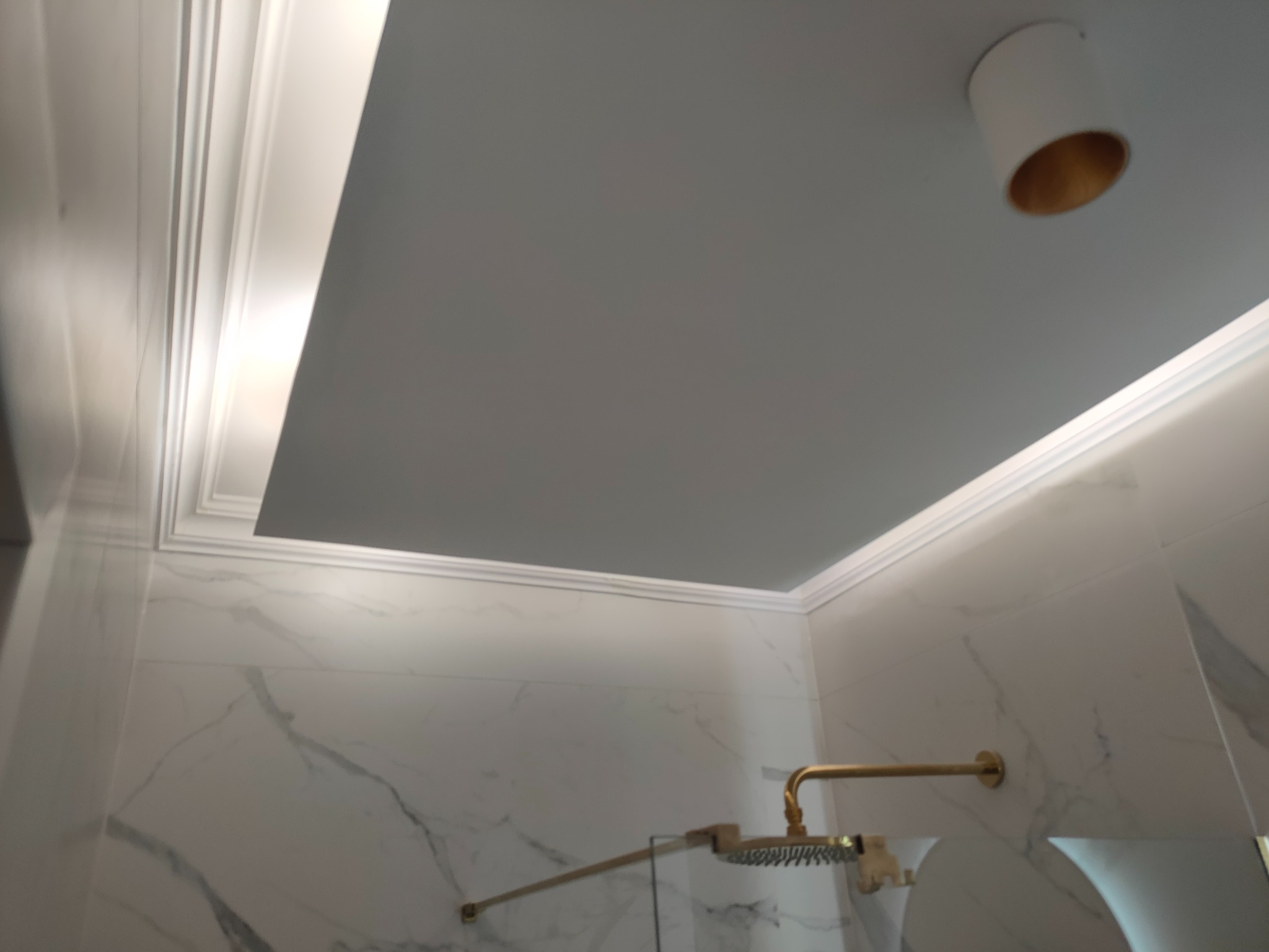 Wykończona łazienka z marmurowymi płytkami, złotą armaturą prysznicową i podwieszanym sufitem z ozdobną sztukaterią i oświetleniem punktowym.