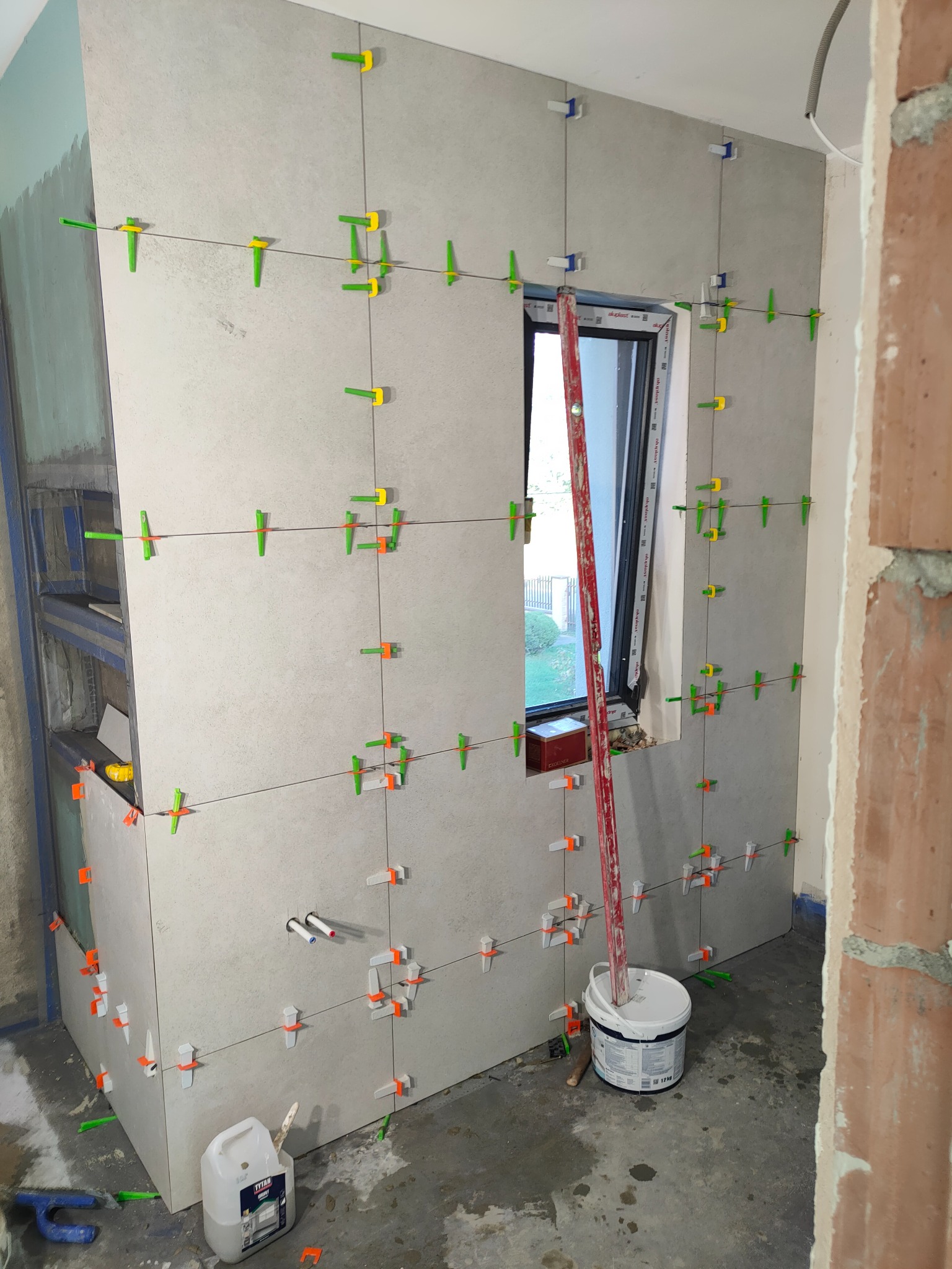 Ściana w trakcie układania płytek ceramicznych w łazience, z widocznym oknem, poziomicą opartą o ścianę, wiadrem z klejem i systemem poziomowania płytek w kolorach zielonym i pomarańczowym.