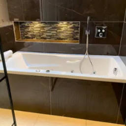 Biała wanna z hydromasażem w obudowie z ciemnych płytek imitujących marmur, z mozaiką w niszy nad wanną, bateria natryskowa zamontowana na ścianie.