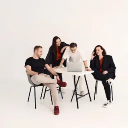 Cztery osoby w studio na białym tle, pracujące przy białym stole z laptopem, kobieta po prawej rozmawia przez telefon, mężczyzna siedzi z założonymi nogami.