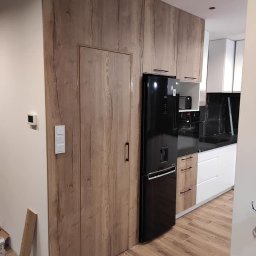 Salon wena Aneta Kępowicz - Nowoczesna kuchnia z zabudową meblową w odcieniach drewna, zintegrowana z drzwiami do pomieszczenia gospodarczego, obok stoi czarna lodówka.