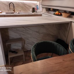 Salon wena Aneta Kępowicz - Nowoczesna kuchnia z wyspą wykończoną marmurem, widoczne krzesła z zieloną tapicerką i drewniany stół. Na wyspie znajduje się otwarta przestrzeń z małym stołkiem.