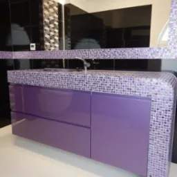 Fioletowa szafka łazienkowa z umywalką, wykończona mozaiką z drobnych płytek, na tle czarnych, błyszczących płytek ściennych.