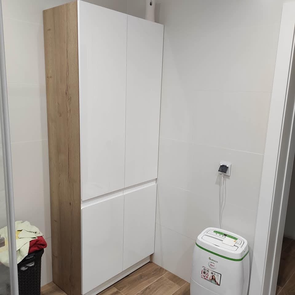 Wysoka szafa z frontami w kolorze białym i bokiem imitującym drewno w łazience z beżowymi płytkami podłogowymi, obok stoi biały osuszacz powietrza.