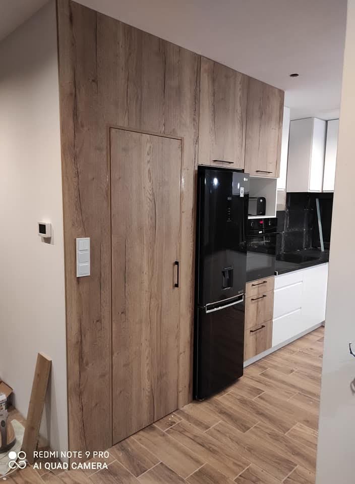 Nowoczesna kuchnia z zabudową meblową w odcieniach drewna, zintegrowana z drzwiami do pomieszczenia gospodarczego, obok stoi czarna lodówka.