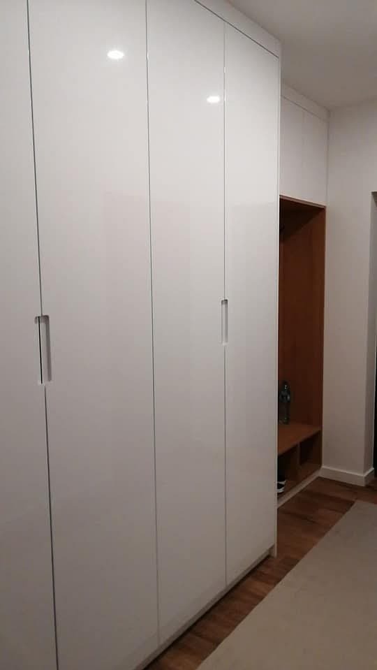 Biała szafa wnękowa z połyskiem, wbudowana w przedpokoju, z widocznym fragmentem drewnianej zabudowy z półkami po prawej stronie.