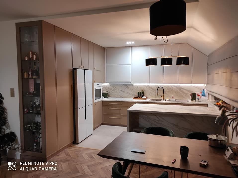 Nowoczesna kuchnia z zabudową w odcieniach beżu i bieli, marmurowym blatem i wyspą, oświetlona trzema czarnymi lampami wiszącymi, widok na stół z czarnymi krzesłami i parkiet w jodełkę.