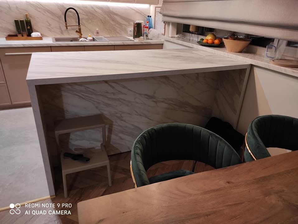 Nowoczesna kuchnia z wyspą wykończoną marmurem, widoczne krzesła z zieloną tapicerką i drewniany stół. Na wyspie znajduje się otwarta przestrzeń z małym stołkiem.