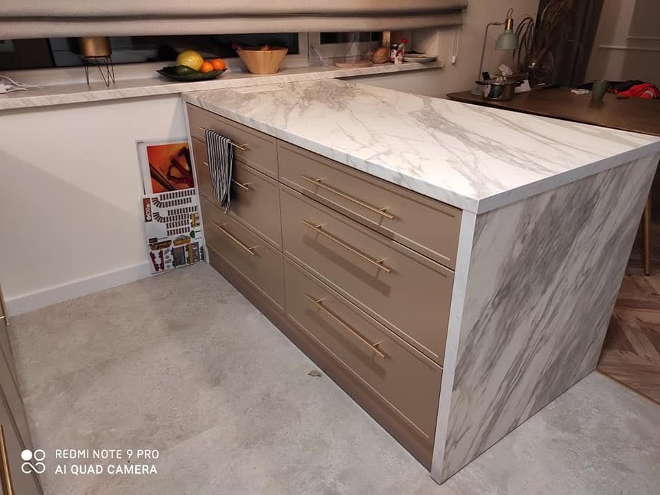 Wyspa kuchenna z beżowymi frontami szuflad i blatem imitującym marmur, złote uchwyty, widoczny fragment podłogi i okna z owocami.