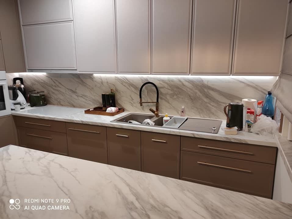 Nowoczesna kuchnia z błyszczącymi, brązowymi szafkami dolnymi i białymi górnymi, marmurowy blat i backsplash, czarna bateria, oświetlenie LED pod szafkami górnymi, widoczny fragment marmurowej wyspy.