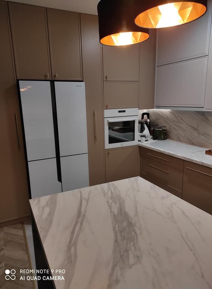 Beżowa zabudowa kuchenna z białym sprzętem AGD i wyspą z blatem imitującym marmur, oświetlona lampami wiszącymi.