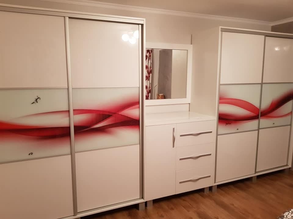 Biała szafa wnękowa z przesuwnymi drzwiami, ozdobiona czerwonym, abstrakcyjnym motywem graficznym, uzupełniona o toaletkę z lustrem i szufladami.