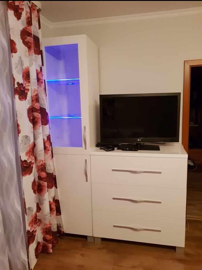 Biała szafka z szufladami i podświetlanymi półkami, na niej stoi telewizor, obok zasłona w kwiaty.