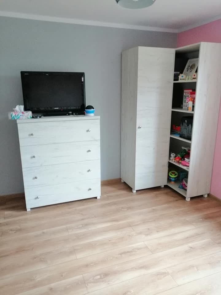 Pokój dziecięcy z białymi meblami: komoda z telewizorem, wysoka szafa z półkami wypełnionymi zabawkami i dekoracjami, panele podłogowe imitujące jasne drewno.