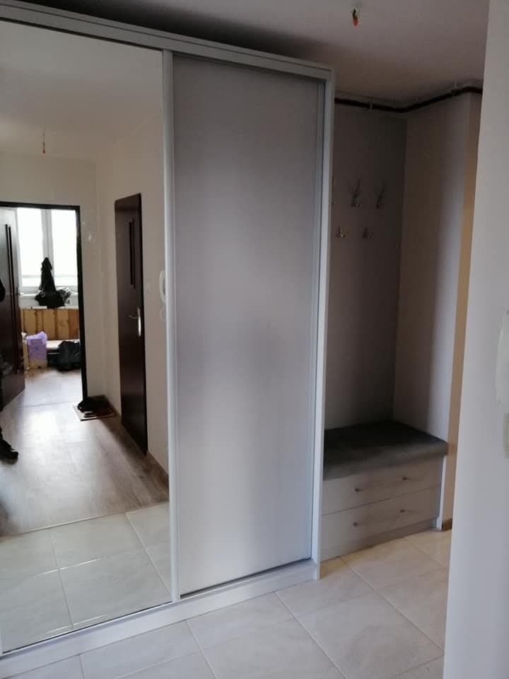 Jasna szafa z przesuwnymi drzwiami z lustrem w przedpokoju, widoczna ławka z szufladami i wieszaki na ścianie.