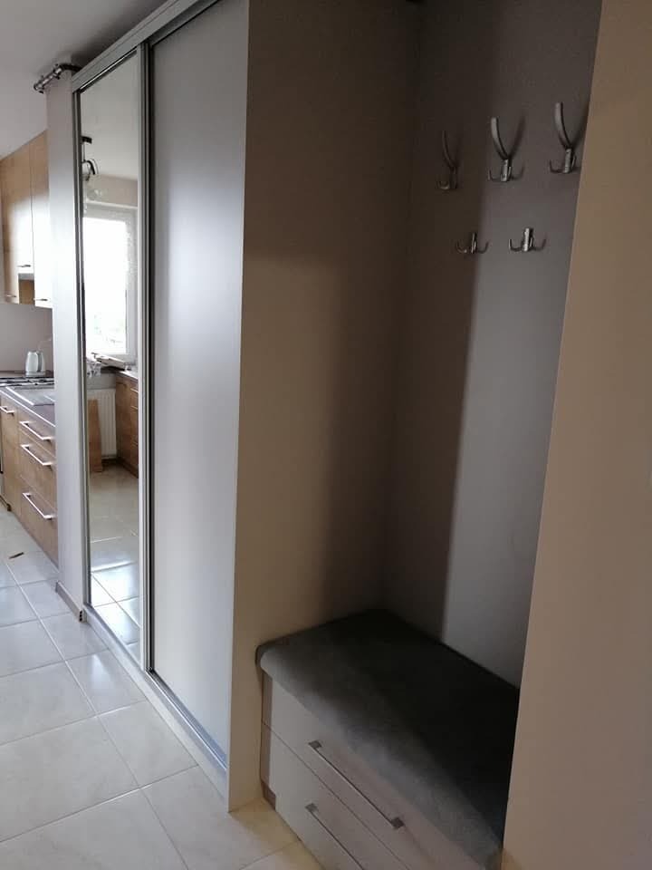 Biała szafa przesuwna z lustrem w przedpokoju, widoczny fragment kuchni w odbiciu, obok siedzisko z szufladami i wieszaki na ścianie.