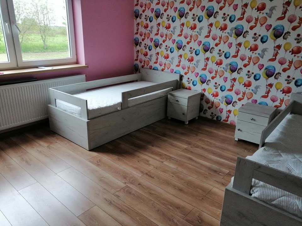 Pokój dziecięcy z drewnianym łóżkiem z barierką, szafką nocną w kolorze jasnego drewna, różową ścianą i tapetą w balony i lisy.