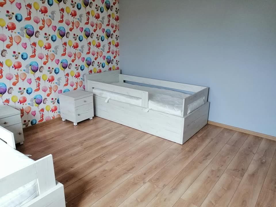 Białe łóżko dziecięce z wysuwanym spaniem i szafka nocna z trzema szufladami na drewnianej podłodze, na ścianie tapeta w balony i zwierzęta.