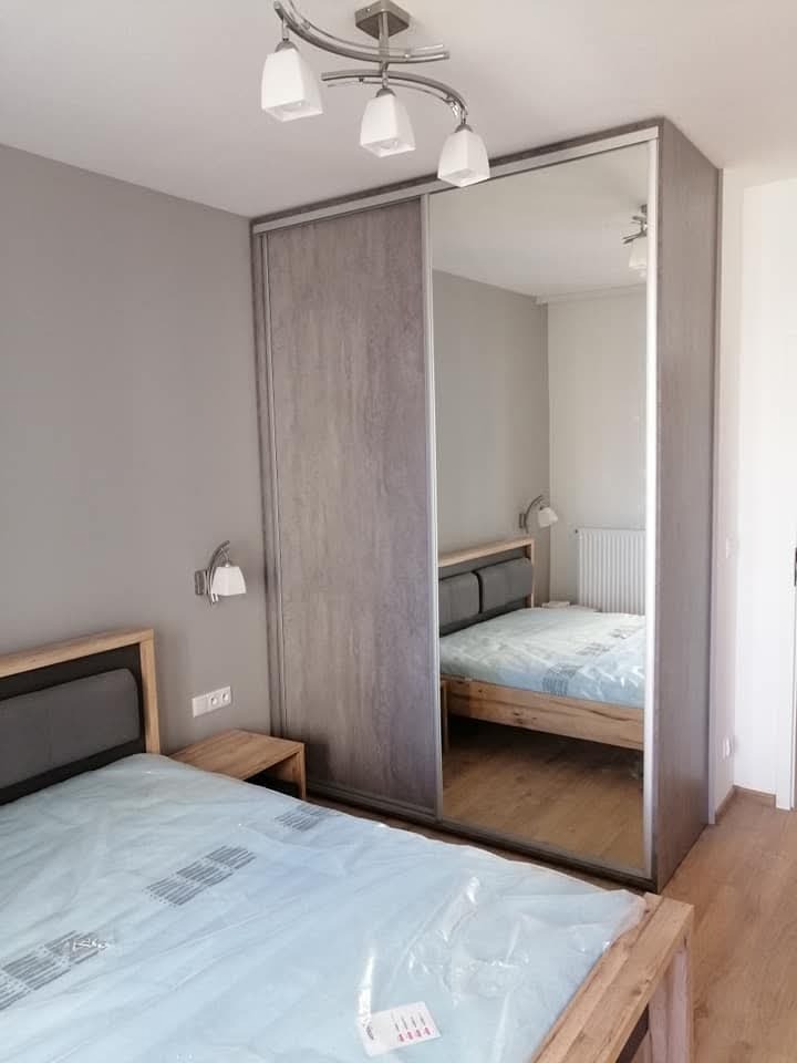 Sypialnia z łóżkiem i szafą wnękową z przesuwnymi drzwiami lustrzanymi; na łóżku folia ochronna.