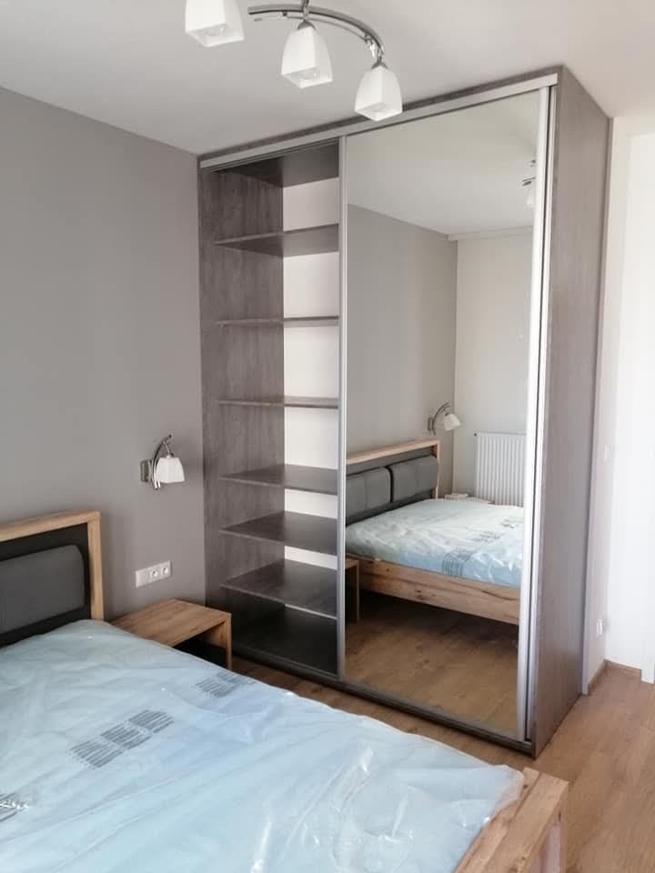 Szara szafa przesuwna z lustrem w sypialni, widoczne półki i odbicie łóżka z zagłówkiem, oświetlenie sufitowe i ścienne.