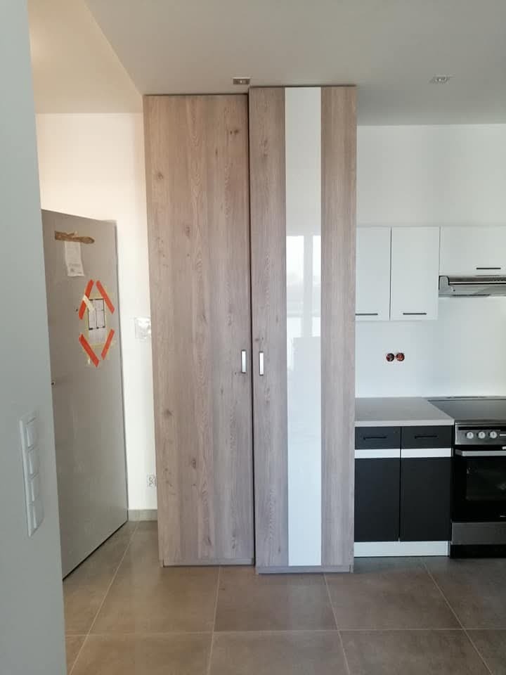 Wysoka zabudowa kuchenna z szafą wnękową o frontach z imitacją drewna i białym pasem, obok białe szafki kuchenne i czarny piekarnik.