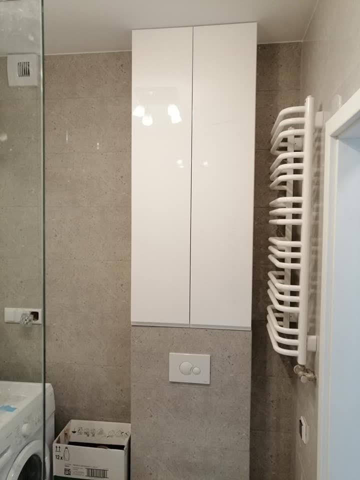 Wysoka, biała szafka łazienkowa z połyskiem, zamontowana nad podtynkową spłuczką, obok grzejnika drabinkowego w łazience wykończonej płytkami w odcieniach szarości.