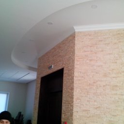 AMGREM SMART BUILDING SOLUTIONS ANATOLII GLADYSH - Wykończenie ściany kamieniem dekoracyjnym wewnątrz pomieszczenia, widoczny fragment sufitu podwieszanego z oświetleniem punktowym oraz ciemne drzwi.