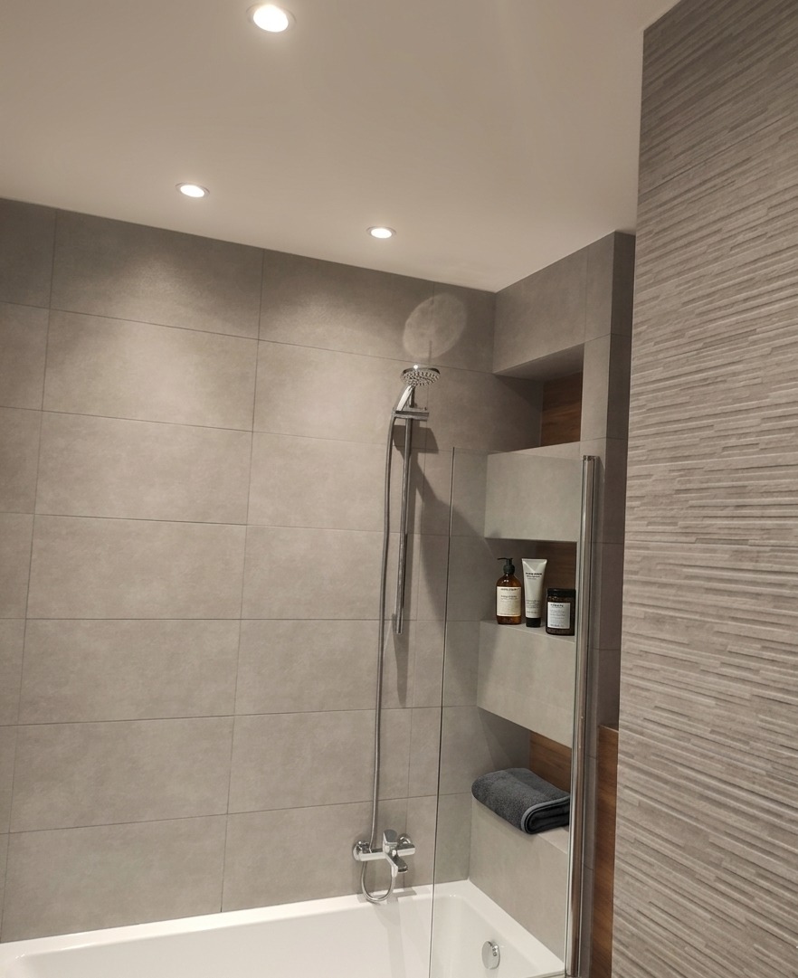 Nowoczesna łazienka z szarymi płytkami, kabiną prysznicową i wnękami na kosmetyki. Minimalistyczny design, oświetlenie punktowe.